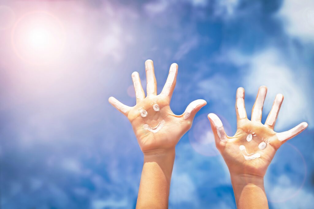 2 hands against the sunny sky with sunscreen smiley faces- Sun Safety blog featured image